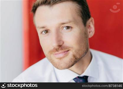 Close up portrait of attractive unshaven male looks at camera, involved in business affairs, being confident in his strength, poses against blurred red and white wall. Man boss with bristle.