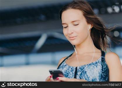 Close up portrait of attractive young woman holds smart phone, texts feedback, checks email box, listens music in player, downloads news songs, connected to wireless internet. People, leisure concept