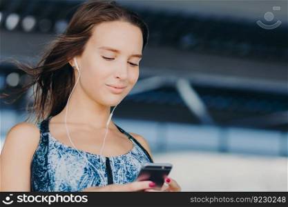 Close up portrait of attractive young woman holds smart phone, texts feedback, checks email box, listens music in player, downloads news songs, connected to wireless internet. People, leisure concept