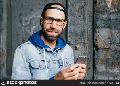 Close-up portrait of bearded blue-eyed man in stylish cap and denim anorak holding mobile phone surfing the Internet cheking his emails and communicating with friends. Technology and communication