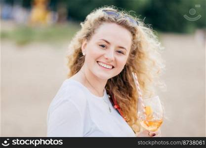 Close up portrait of beautiful European female with light wavy hair, toothy smile, holds cold fresh beverage, enjoys sunny good weather, poses against blurred beach background. People and drink