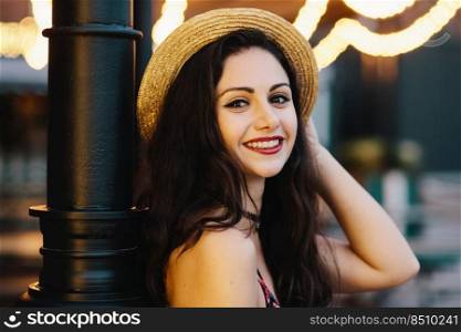 Close up portrait of beautiful female with dark appealing eyes, smiling gently at camera while posing indoors at cafeteria. Romantic young attractive woman with make-up wearing straw summer hat