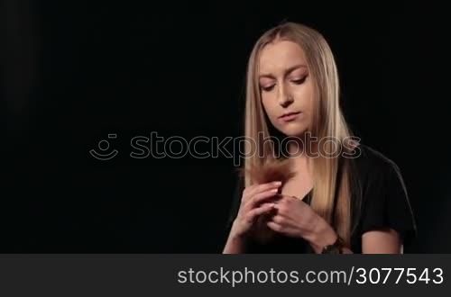 Close up portrait of beautiful frustrated young girl because of tangled ends of her long blond hair on black. Beautiful woman holding split tips of her amazing straight hair and looking at camera full of sadness. Beauty hairstyle concept.