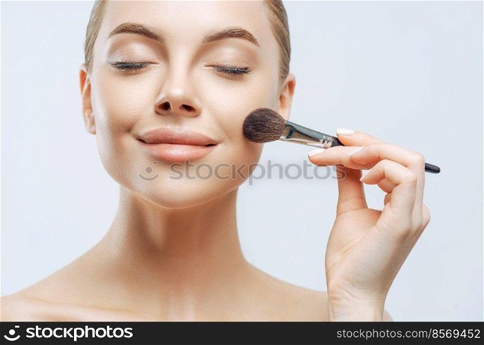 Close up portrait of beautiful girl has fresh healthy skin, closes eyes, applies cosmetics on face, uses beauty brush, has professional makeup, poses indoor. Cosmetology, visage, wellness concept