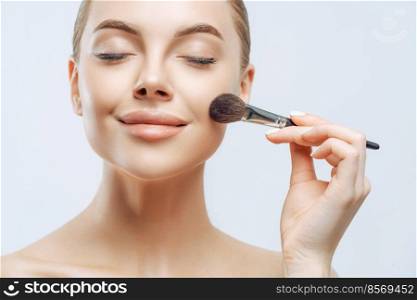 Close up portrait of beautiful girl has fresh healthy skin, closes eyes, applies cosmetics on face, uses beauty brush, has professional makeup, poses indoor. Cosmetology, visage, wellness concept