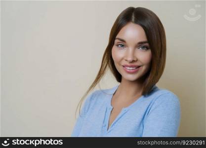 Close up portrait of beautiful lady with beauty face and healthy clean skin, smiles pleasantly, has shiny straight brown hair, looks confidenlty at camera, isolated over beige wall. Hair product, care