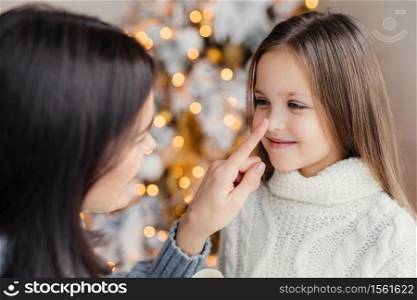 Close up portrait of beautiful little girl with long hair, has fun with her mother, looks in eyes, stand together against decorated fir tree with garlands and lights background. Celebration concept