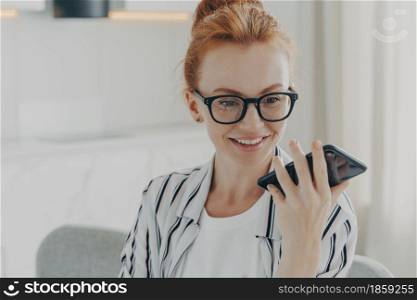 Close-up portrait of beautiful positive red-haired woman in spectacles holding mobile phone and sending audio message to friend while sitting in whote kitchen at home, using speakerphone loudspeaker. Beautiful positive red-haired woman in spectacles holding mobile phone and sending audio message