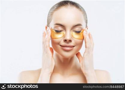 Close up portrait of beautiful smiling European woman closes eyes, enjoys anti aging eye therapy, applies golden patches, has fresh clean skin, stands shirtless against white background. Face care