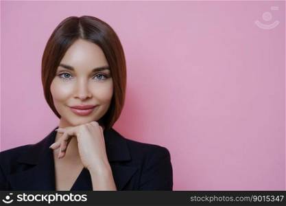 Close up portrait of beautiful woman keeps hand under chin, looks directly at camera, has blue eyes and full lips, listens attentively interlocutor, wears formal outfit, isolated on pink background