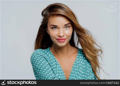 Close up portrait of beautiful young female has makeup, dressed in polkadot shirt, works as fashion model poses against white background, wears lipstick, has healthy smooth skin. Facial expressions