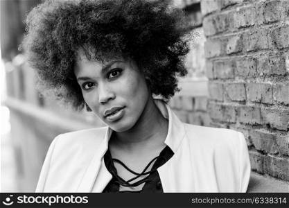 Close-up portrait of black woman with afro hairstyle standing in urban background. Mixed girl wearing white jacket and black dress posing near a brick wall