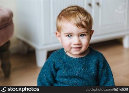 Close up portrait of blonde little child with plump cheeks, looks with blue eyes into camera, has innocent look. Adorable infant has serious expression sits over home interior. Childhood concept