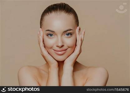 Close up portrait of blue eyed glad woman with dark combed hair, touches face gently, has healthy perfect skin, bare shoulders, enjoys beauty procedures, models indoor. Skin care and wellness