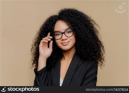 Close up portrait of calm satisfied Afro American woman with bushy curly hairstyle, keeps hand raised, has healthy skin, wears spectacles, black elegant outfit isolated on brown wall. Face expressions