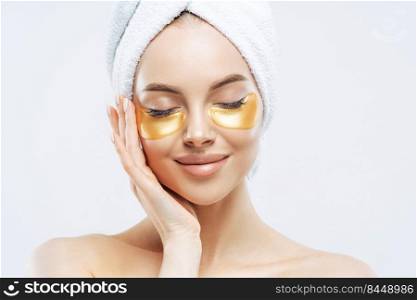 Close up portrait of charming young woman with cosmetic collagen patches under eyes, enjoys flawless of skin, has well cared complexion, manicure, wears wrapped towel on head, stands indoor.