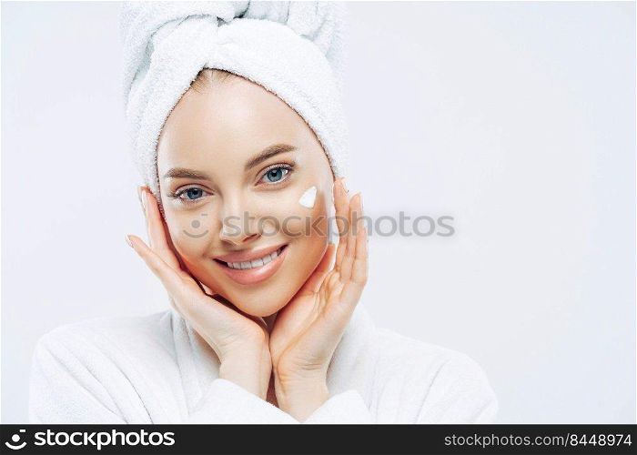 Close up portrait of cheerful female with natural beauty, touches cheeks gently, applies face cream for soft skin, wears bath towel on head, dressed in robe, stands indoor. Beauty treatment, lifestyle
