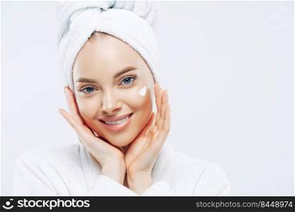 Close up portrait of cheerful female with natural beauty, touches cheeks gently, applies face cream for soft skin, wears bath towel on head, dressed in robe, stands indoor. Beauty treatment, lifestyle