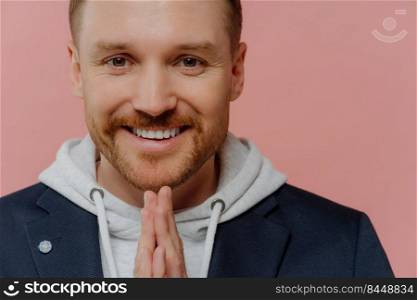 Close up portrait of cheerful redhead bearded man in casual clothes holding hands in praying gesture above chin and asking for good luck, hoping or making wish while standing over pink wall. Happy man making praying gesture and smiling at camera
