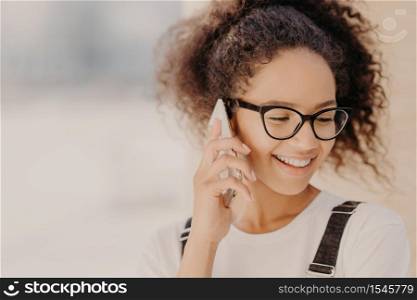Close up portrait of cheerful woman with crisp hair, satisfied with tariffs for telephone call, focused down, holds modern smart phone, phones to best friend, discusses latest news. Technology concept