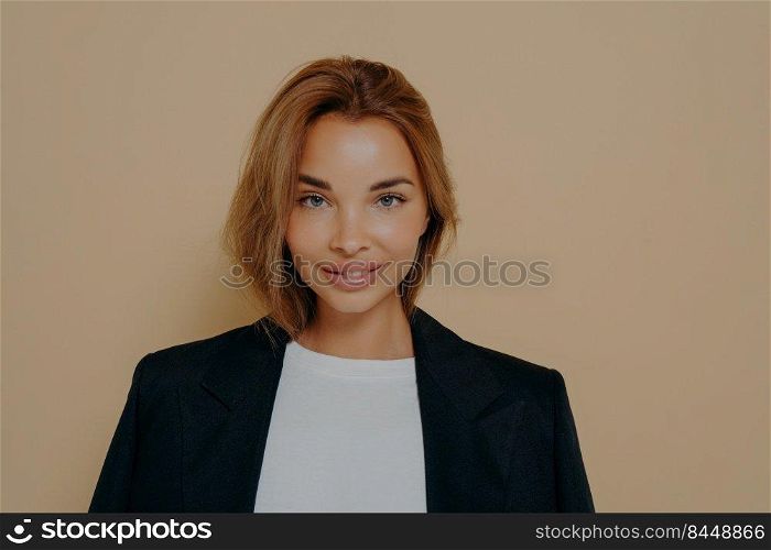 Close up portrait of confident smiling business woman in formal black blazer worn over white blouse, posing in studio isolated against beige background with blank space.Young female model. Close up portrait of confident smiling business woman in formal black blazer