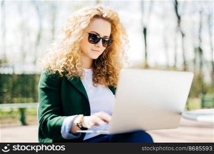 Close-up portrait of cute female with fluffy blonde hair and pure skin wearing sunglasses and green jacket concentrated on screen of her laptop reading attentively interesting stories sitting at bench