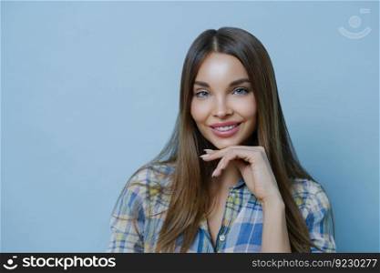 Close up portrait of cute friendly looking lady touches chin, has healthy skin, minimal make up, looks with warm tender expression, has well cared hair poses against blue background. Beauty, skin care