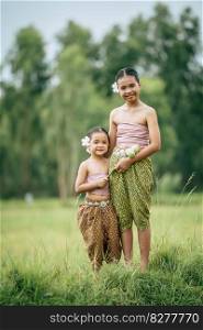 Close up, Portrait of Cute sister and young sister in Thai traditional dress and put white flower on her ear standing in rice field,  smile and looking at camera, sibling love concept, copy space