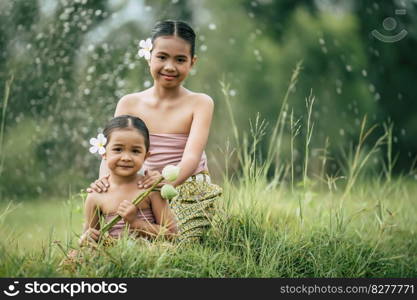 Close up, Portrait of Cute sister and young sister in Thai traditional dress and put white flower on her ear sitting in meadow,  smile and looking at camera, sibling love concept, copy space