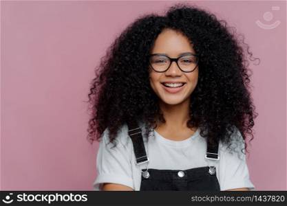 Close up portrait of dark skinned woman with crisp hair, smiles happily, wears optical glasses, casual clothes, poses over purple background expresses good emotions. Afro American student poses indoor