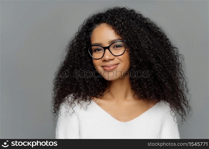 Close up portrait of delighted beautiful Afro woman with bushy curly hair, looks through transparent glasses, wears white sweater, poses against grey studio wall. Ethnicity, beauty, facial expressions