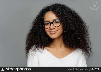 Close up portrait of delighted beautiful Afro woman with bushy curly hair, looks through transparent glasses, wears white sweater, poses against grey studio wall. Ethnicity, beauty, facial expressions