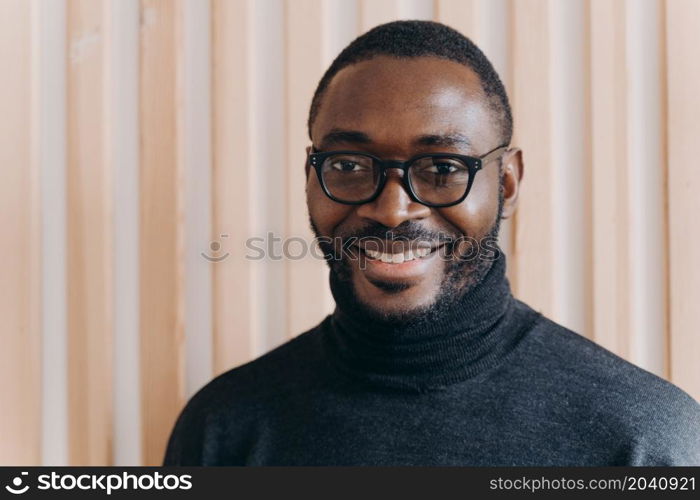 Close up portrait of elegant confident African american businessman in glasses posing while standing in modern office, looking at camera smiling broadly and sincerely, happy with company progress. Elegant confident Afro american businessman in glasses posing while standing in modern office