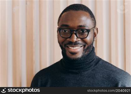Close up portrait of elegant confident African american businessman in glasses posing while standing in modern office, looking at camera smiling broadly and sincerely, happy with company progress. Elegant confident Afro american businessman in glasses posing while standing in modern office