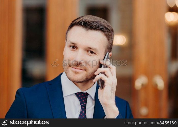 Close up portrait of good looking male with attractive appearance, speaks via cell phone, manages financial problems with colleague, stands outdoor, dressed in formal clothing. Communication