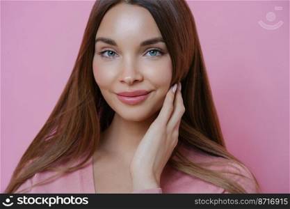 Close up portrait of gorgeous European woman with healthy perfect skin, touches her well cared hair, looks directly at camera with blue eyes, poses against pink background. Natural beauty concept