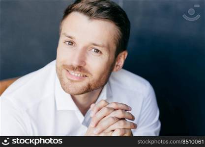 Close up portrait of handsome unshaven male with positive smile, keeps hands together, feels successful as works in business sphere, wears white shirt, isolated over dark background. Emotions concept