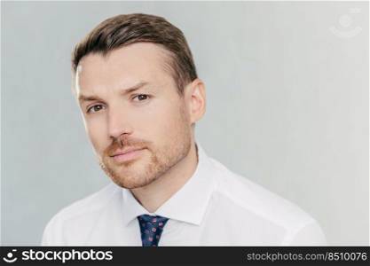 Close up portrait of handsome unshaven man looks seriously at camera, has appealing look, thinks about business affairs, dressed in formal clothes, isolated over white background. CEO indoor
