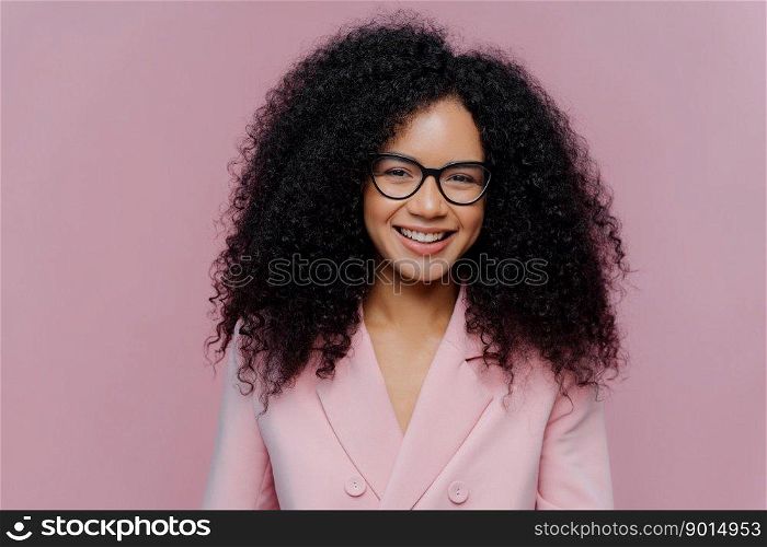 Close up portrait of happy curly haired woman has gentle smile on face, dark healthy skin bushy hair, wears elegant clothes, isolated on purple background, enjoys coming weekend, works in office