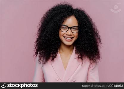 Close up portrait of happy curly haired woman has gentle smile on face, dark healthy skin bushy hair, wears elegant clothes, isolated on purple background, enjoys coming weekend, works in office