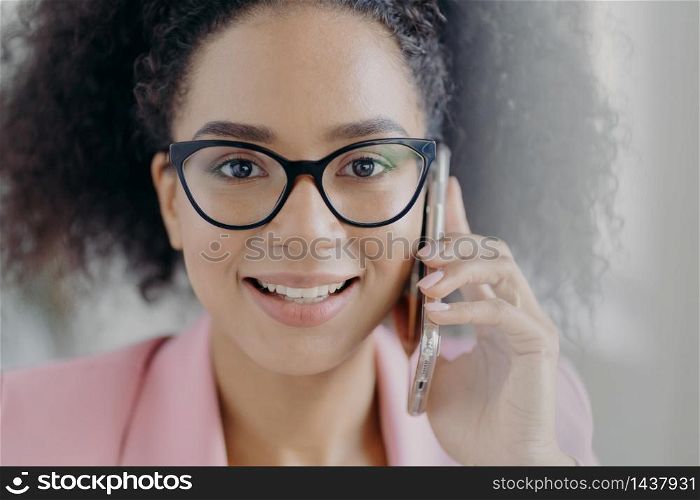 Close up portrait of happy dark skinned businesswoman holds mobile phone near ear, calls business partner, has charming smile, wears optical glasses, has curly bushy hair, discusses working issues