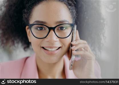 Close up portrait of happy dark skinned businesswoman holds mobile phone near ear, calls business partner, has charming smile, wears optical glasses, has curly bushy hair, discusses working issues