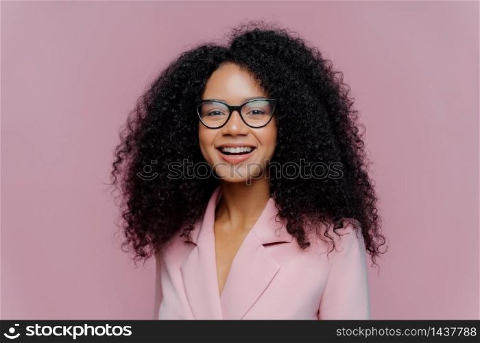 Close up portrait of happy ethnic young woman with crisp hair, looks through optical glasses, wears formal outfit, being in good mood, comes on work, happy get praise from boss, models indoor