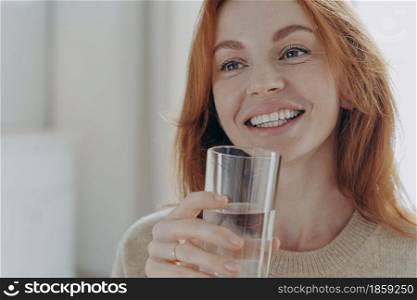 Close up portrait of happy healthy ginger woman holding transparent glass of pure mineral water, smiling cheerfully and enjoying new day while standing in morning at home, taking care about health. Portrait of happy healthy ginger woman holding transparent glass of pure mineral water