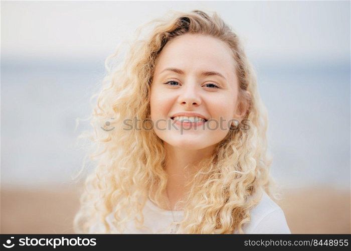 Close up portrait of happy young cute female with curly hair, looks gladfully directly at camera, has broad smile, poses against seaside background. People, beauty and positive emotions concept