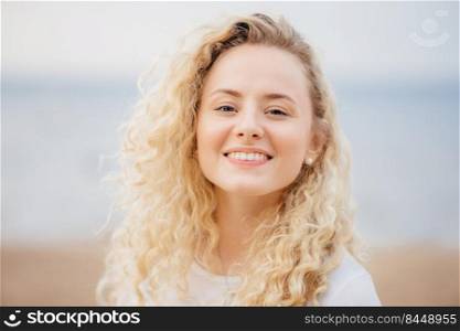 Close up portrait of happy young cute female with curly hair, looks gladfully directly at camera, has broad smile, poses against seaside background. People, beauty and positive emotions concept