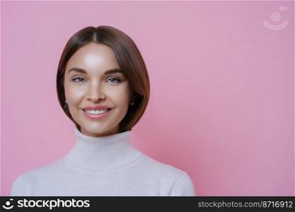 Close up portrait of lovely satisfied joyful woman with dark hair, smiles pleasantly, shows white teeth, wears poloneck, isolated on pink background, empty space for your advertising content.