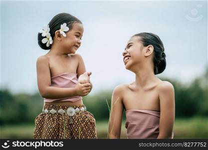 Close up, Portrait of lovely sister and young sister in Thai traditional dress and put white flower on her ear, Look into each other’s eyes and laugh happily on green nature background, copy space