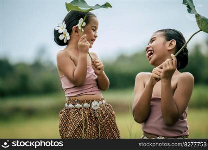Close up, Portrait of lovely sister and young sister in Thai traditional dress and put white flower on her ear, Look∫o each other’s eyes and laugh hapπly on green nature background,©space