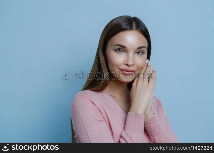 Close up portrait of pleased attractive brunette woman gazes directly at camera, keeps hands near face, listens advice from cosmetologist how to care about skin, models indoor against blue wall
